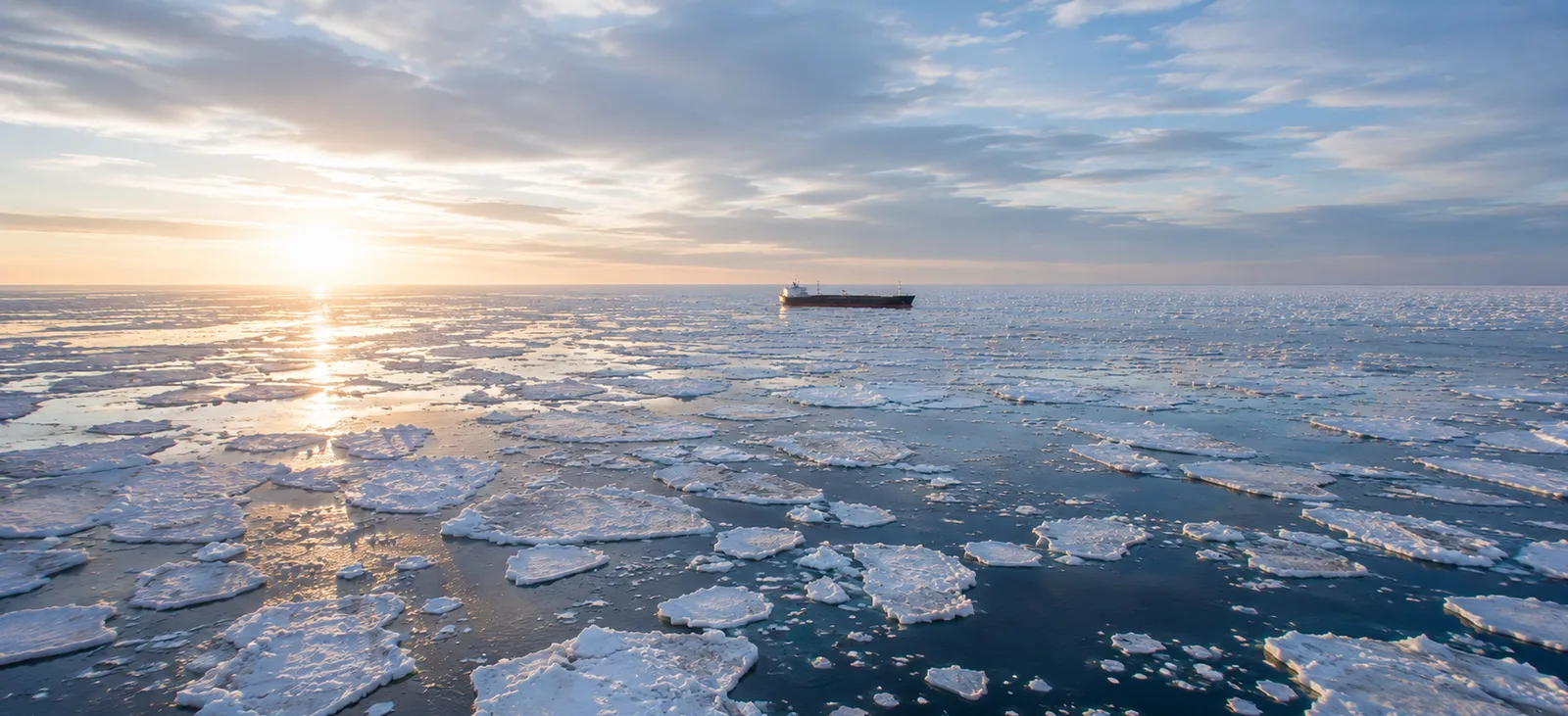 Elevated view of breaking spring pack ice melting in dark blue Arctic water with a distant cargo ship in the middle distance, suggesting seasonal route opening