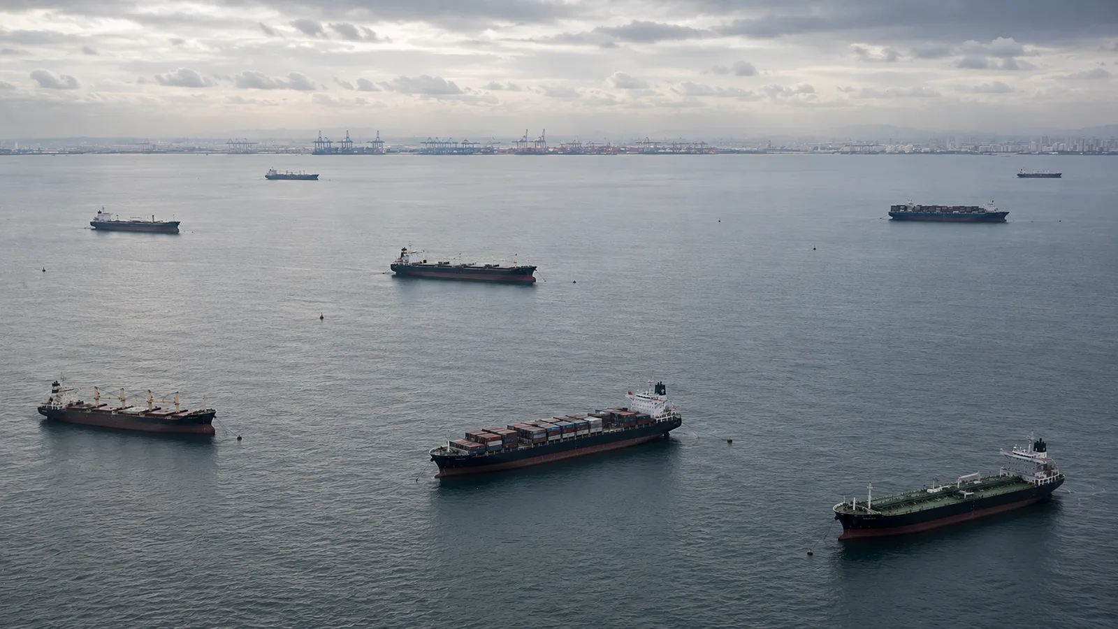 Aerial overhead view of a busy port anchorage with multiple commercial vessels at anchor in loose formation — container ships, bulk carriers, product tankers