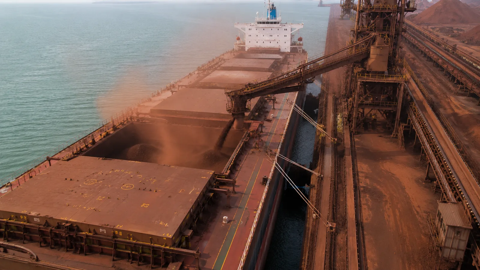 Bulk carrier loading iron ore at a terminal — conveyor system and open hatch visible