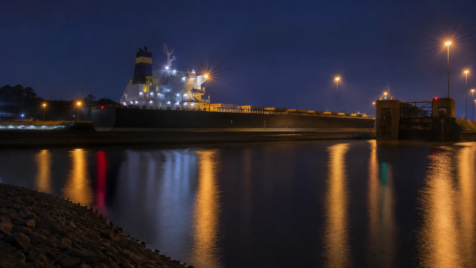 A cargo ship in a canal lock at night, deck floodlights reflecting on still water beside the lock gates